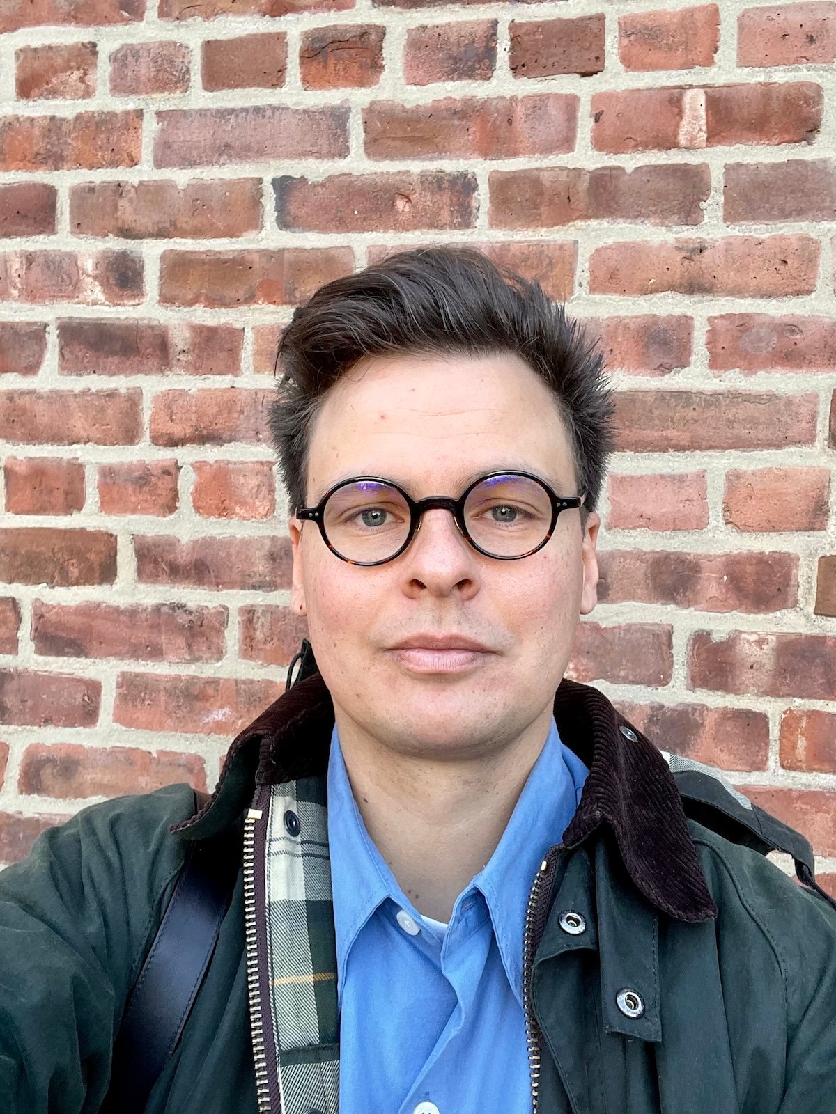 A photograph of a man with brown hair and dark-framed glasses wearing a blue buttondown shirt and a green work coat.