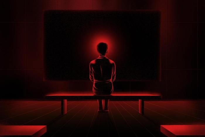 The back of a female figure sitting on a bench, with a red halo around her head, against a background of a dark projection screen.